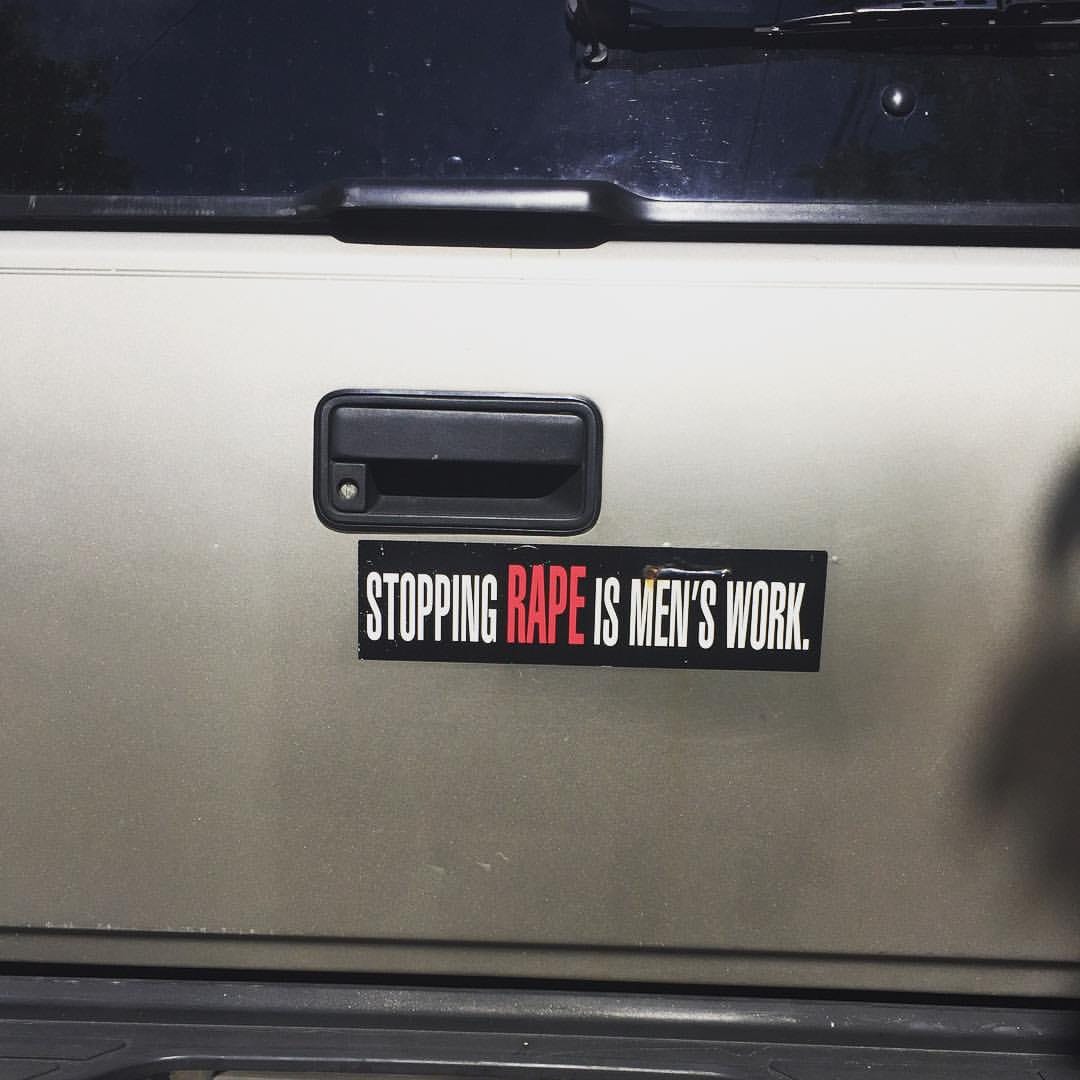 A bumper sticker on a vehicle says "STOPPING RAPE IS MEN'S WORK." The words are white except for "rape" which is red and enlarged.