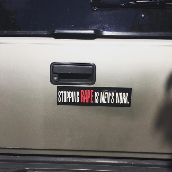 A bumper sticker on a vehicle says "STOPPING RAPE IS MEN'S WORK." The words are white except for "rape" which is red and enlarged.